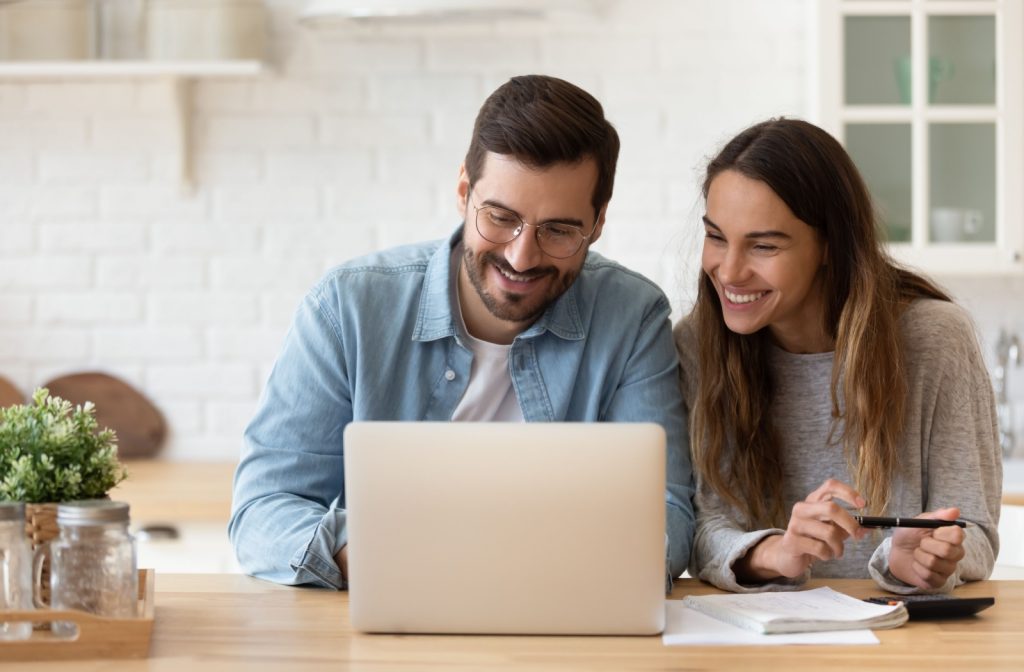A young couple happily look over their laptop in a brightly lit kitchen organizing their finances.